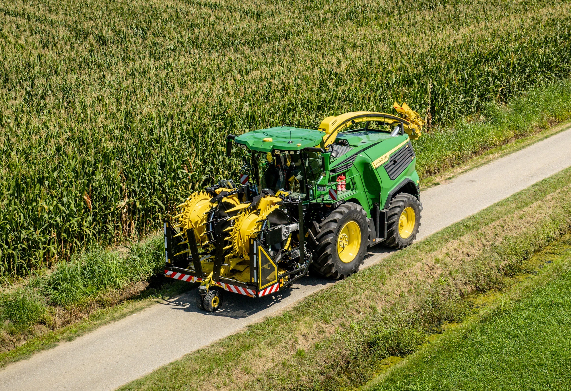 Μπροστινή αριστερή όψη μιας αναδιπλωμένης κεφαλής κοπής σε αυτοκινούμενο ενσιρωκοπτικό μηχάνημα που κινείται παράλληλα με ένα χωράφι καλαμποκιού.