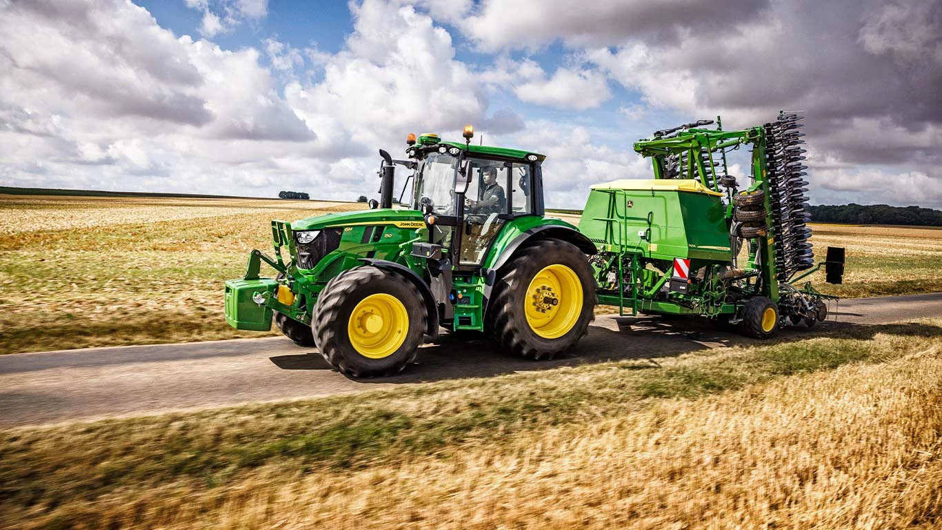 Ένα τρακτέρ που ρυμουλκεί μια ρυμούλκα κινείται σε αγροτικό δρόμο, ο οποίος περιβάλλεται από πράσινο και ανοιχτούς αγρούς.