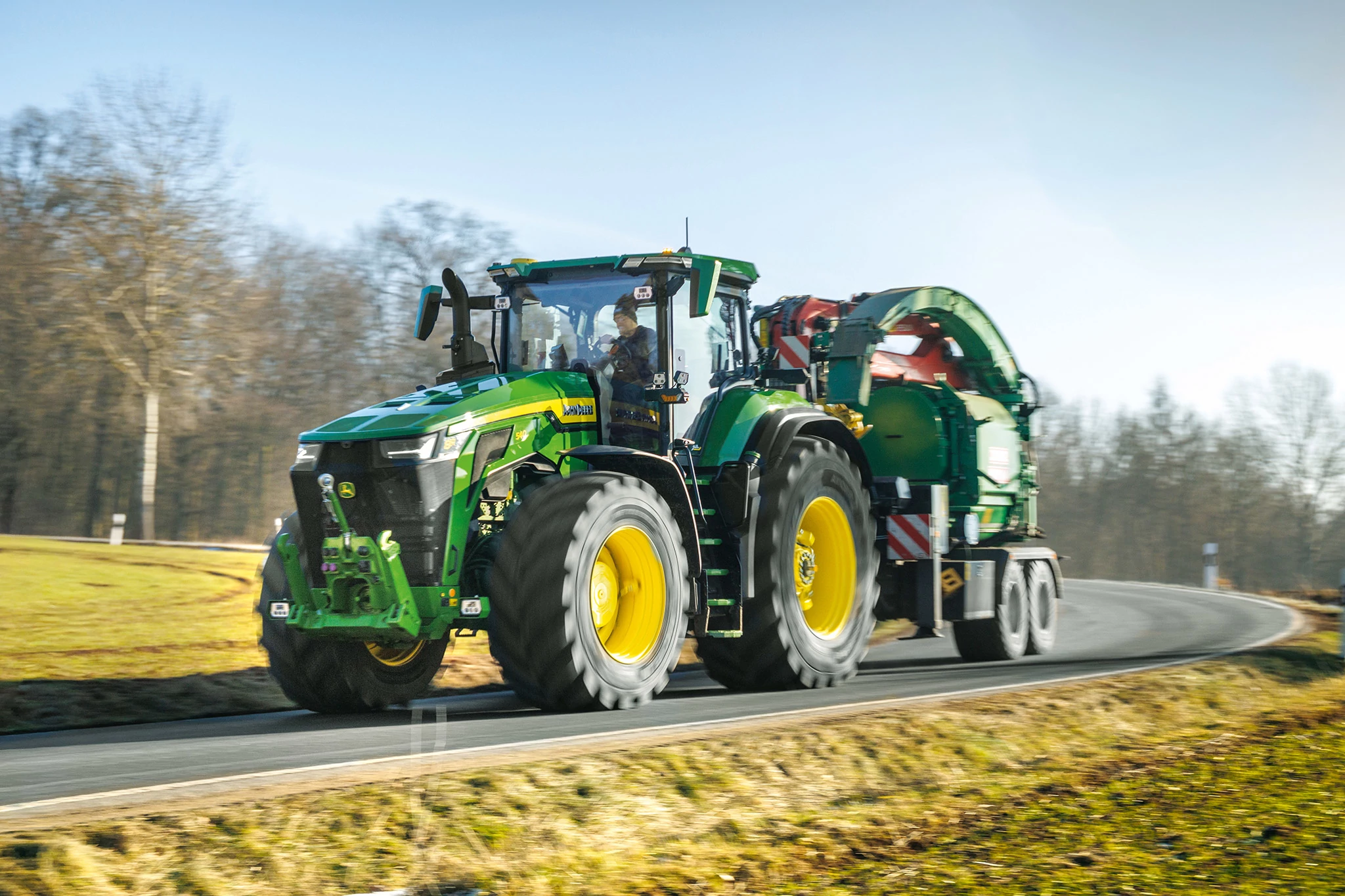 Τρακτέρ 8R 540 που κινείται σε ασφαλτοστρωμένο δρόμο, ενώ ρυμουλκεί ένα μεγάλο πράσινο παρελκόμενο, με λιβάδια και δέντρα στο φόντο