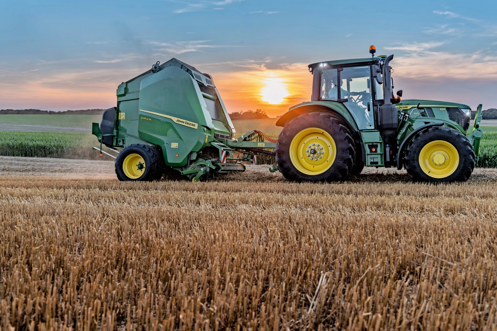 John Deere V462M σε έναν αγρό άχυρου που ρυμουλκείται από ένα John Deere 6M 150 κατά τη διάρκεια του ηλιοβασιλέματος