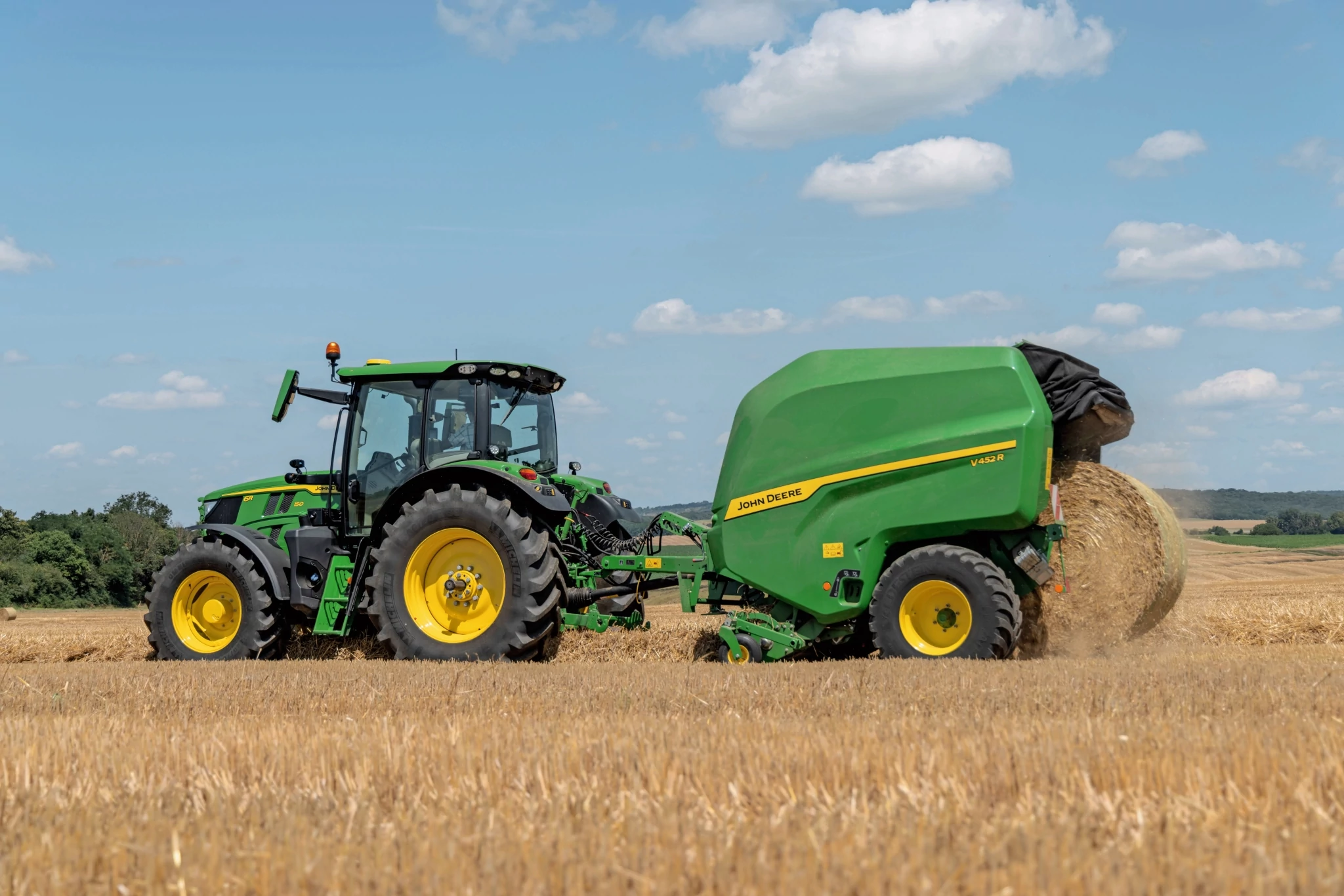 Χορτοδετικό μεταβλητού θαλάμου John Deere σε έναν αγρό άχυρου που ρυμουλκείται από ένα John Deere 6R 150 που εξάγει ένα δέμα