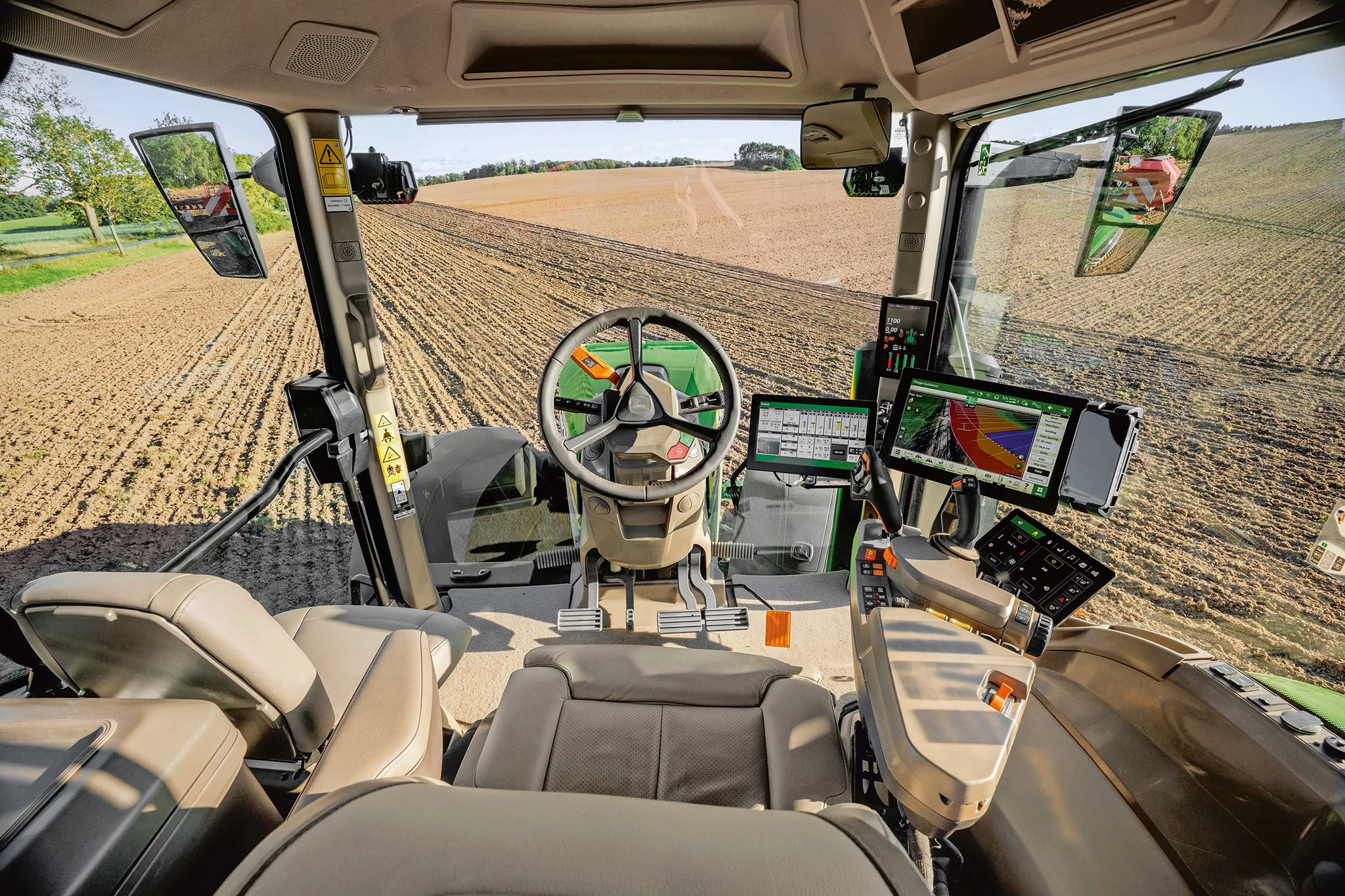 Η εσωτερική όψη μιας σύγχρονης καμπίνας τρακτέρ εμφανίζει το κάθισμα χειριστή, το τιμόνι, την κονσόλα ελέγχου και πολλές ψηφιακές οθόνες. Τα μεγάλα παράθυρα παρέχουν μια ευρεία θέα ενός φρεσκοκαλλιεργούμενου αγρού έξω.