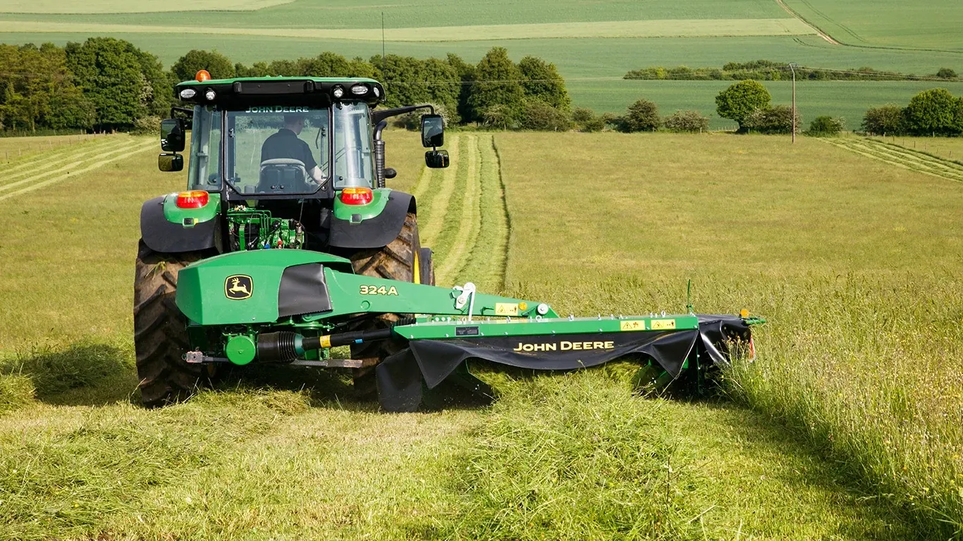 Χλοοκοπτικό με δίσκο πίσω στερέωσης John Deere 324A που κόβει χόρτο σε αγρό, απεικονίζεται από πίσω με το τρακτέρ σε επικλινές έδαφος.