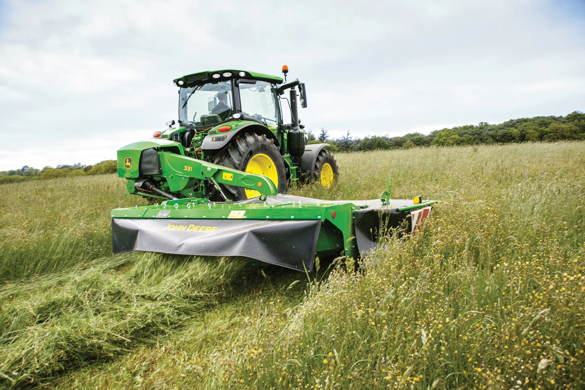 Το χορτοκοπτικό-δίσκος πίσω στερέωσης John Deere 331 κόβει ψηλό χόρτο σε έναν αγρό, όπως φαίνεται από τη γωνιακή πίσω όψη με το τρακτέρ.