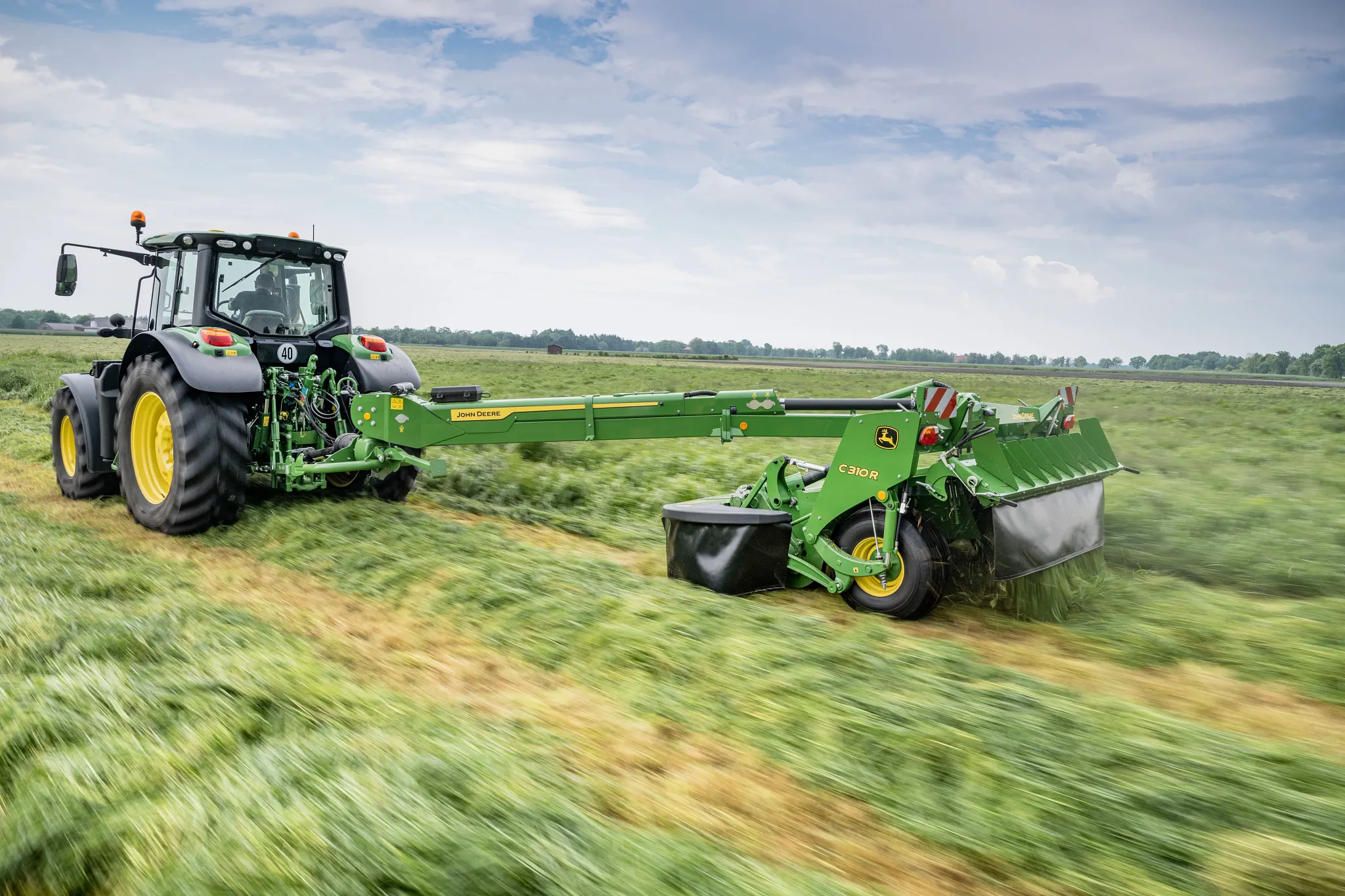Ρυμουλκούμενο χορτοκοπτικό John Deere 310R που κόβει χόρτο στον αγρό, απεικονίζεται από την γωνιακή πίσω πλευρά με το τρακτέρ να τραβάει το μηχάνημα.