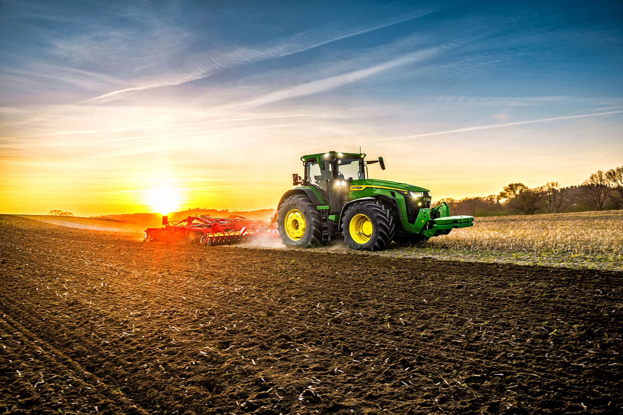 Tractor cultivating field at sunset, John Deere 8R pulling cultivator across prepared soil