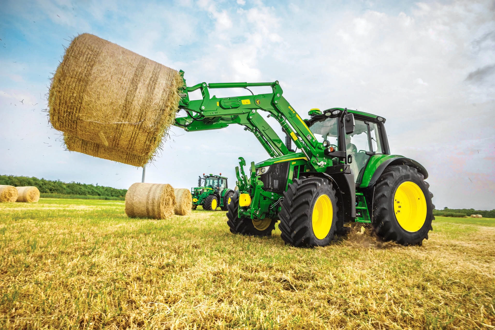 Τρακτέρ σειράς 6M της John Deere που ανυψώνει ένα μεγάλο στρογγυλό δέμα σανού με μπροστινό φορτωτή σε ανοιχτό χωράφι.