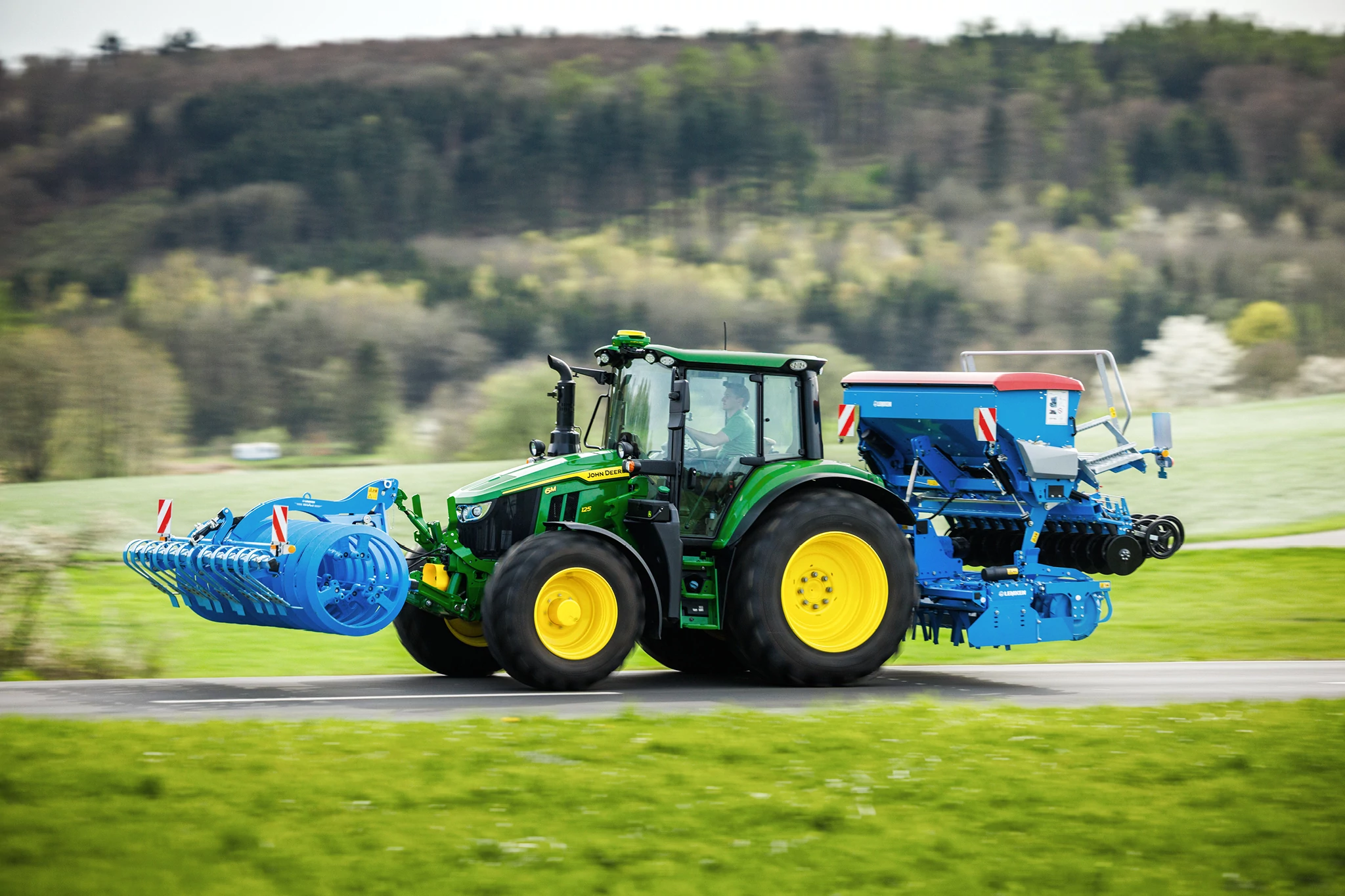 John Deere 6M Series tractor transporting a front-mounted implement and rear seeder along a rural road.